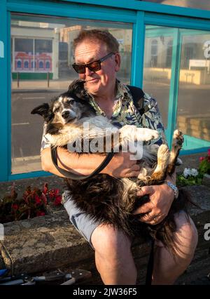 Ein Mann kuschelt seinen älteren walisischen Collie-Schafhund am Meer in Worthing, West Sussex, Großbritannien. Stockfoto