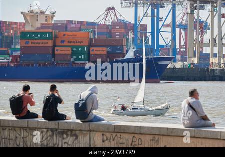 03. September 2022, Hamburg: Die Menschen genießen die Sonne und den Blick auf den Containerhafen am Elbestrand in Övelgönne, während ein Segelboot stromabwärts fährt. Foto: Georg Wendt/dpa Stockfoto