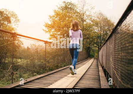 Fitness und sportliche Motivation. Rückansicht einer sportlichen Läuferin, die an einem kühlen Herbsttag über eine alte Hängebrücke läuft. Cardio-Training im Freien Stockfoto