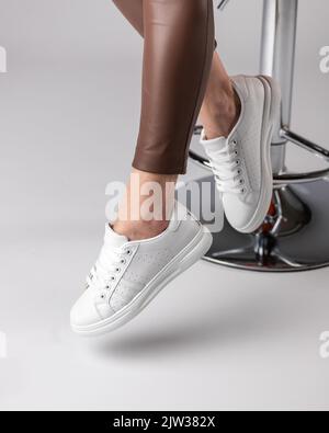 Stilvolle moderne Damen-Sneakers auf den Beinen des Models, das auf einem Stuhl im Studio auf weißem Hintergrund sitzt. Sneakers sind sehr beliebt bei Teenagern oder Frauen Stockfoto