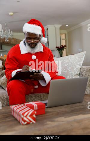 Älterer afroamerikanischer Mann, der ein Kostüm des weihnachtsmannes trägt und auf der Zwischenablage schreibt Stockfoto