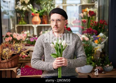 Attraktiver junger Kaukasier mit Brillen und Beanie im Blumengeschäft mit Tulpenstrauß. Gut aussehende männliche Floristin in Pullover hält Strauß. Konzept für kleine Unternehmen. Hochwertige Bilder Stockfoto