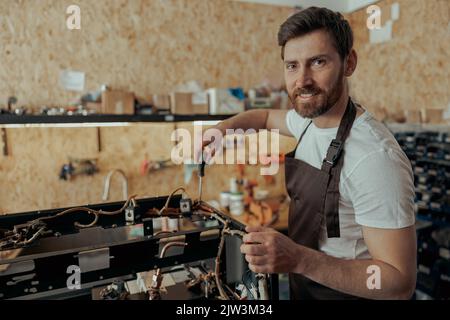 Lächelnder junger Mann, der die Kaffeemaschine mit einem Schraubendreher in einer Werkstatt und einer aussehenden Kamera repariert Stockfoto