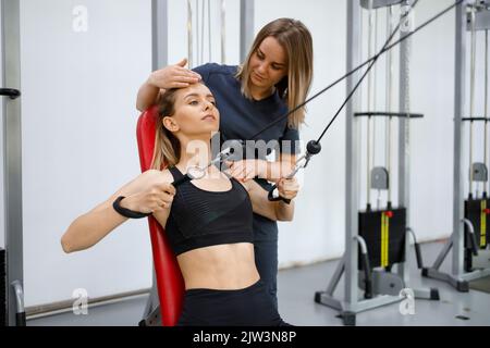 Junge Patientin im Fitnessstudio, die unter der Aufsicht eines Reha-Arztes körperliche Übungen auf dem Weichen-Kabelgerät macht Stockfoto