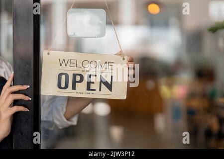 Inhaber eines Kleinunternehmens lächelt, während er das Schild für die Öffnung des Ortes nach der Quarantäne wegen Covid-19 dreht. Nahaufnahme der Frau, die die Hände hält Stockfoto