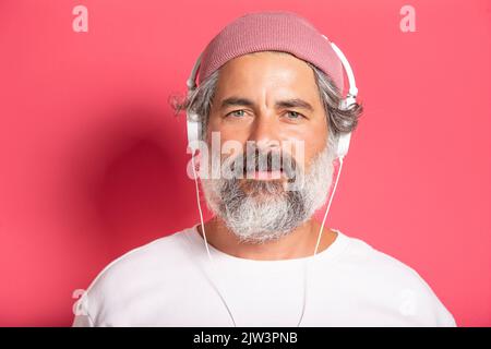 Nahaufnahme eines hübschen graubärtigen Mannes in Ohrhörern, der Musik hört oder mit Kopfhörern auf dem Mobiltelefon spricht, fröhlich lächelnd auf die Kamera lächelt und im Stehen steht Stockfoto