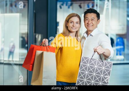 Junge, vielfältige Familie, asiatischer Mann und blonde Frau, die im Supermarkt einkaufen, Porträt von ein paar Käufern, die die Kamera betrachten und lächeln und bunte Taschen mit Waren und Einkaufen halten. Stockfoto