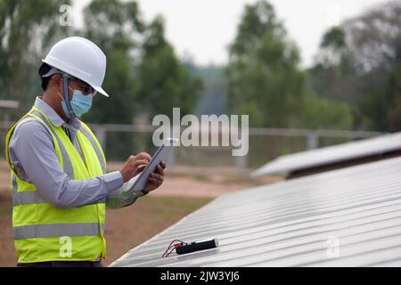Ein junger asiatischer Ingenieur, der einen Helm und eine weiße Maske trägt, überprüft die Sauberkeit von Solarmodulen. Konzept für erneuerbare Energien Stockfoto