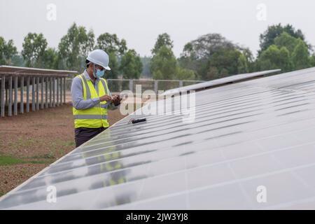 Ein junger asiatischer Ingenieur, der einen Helm und eine weiße Maske trägt, überprüft die Sauberkeit von Solarmodulen. Konzept für erneuerbare Energien Stockfoto