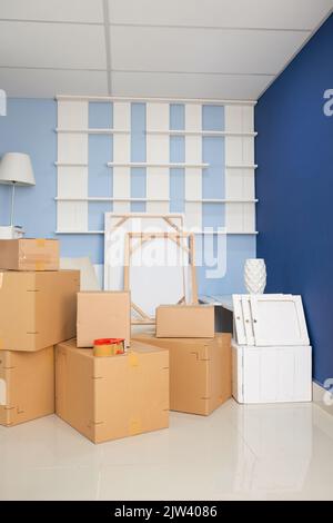 Gepackte Pappkartons mit Haushaltswaren, die auf dem Boden in einem leeren Raum mit blauen Wänden gestapelt wurden Stockfoto