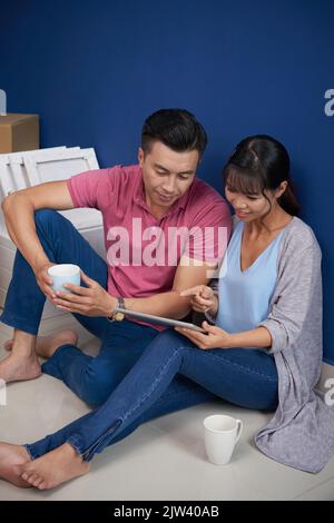 Junges asiatisches Paar, das in einer neuen Wohnung auf dem Boden sitzt, sich mit Tassen Kaffee ausruhte und ein digitales Tablet verwendete Stockfoto