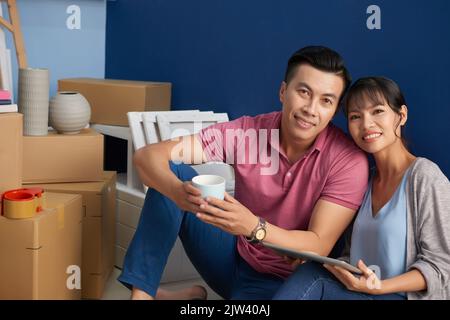 Porträt eines jungen asiatischen Paares, das fröhlich auf die Kamera lächelt, während es in einer neuen Wohnung neben verpackten Kartons und Haushaltsgegenstand sitzt Stockfoto