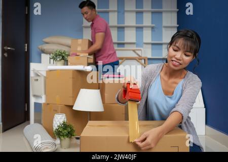 Junges asiatisches Paar, das Sachen zu Hause verpackt, während es in neues Haus umzieht, Frau, die Pappkarton mit Klebeband verpackt Stockfoto