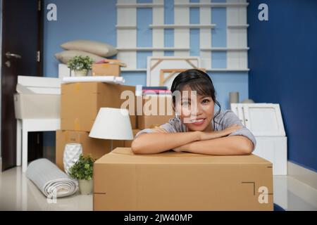 Porträt einer jungen, schönen asiatischen Frau, die sich auf einen Karton lehnte und die Kamera anlächelte, während sie in einer neuen Wohnung auspackte Stockfoto