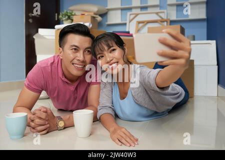 Junges asiatisches Paar, das Selfie auf dem Smartphone nimmt und glücklich lächelt, während es in einer neuen Wohnung mit verpackten Boxen auf dem Boden liegt Stockfoto
