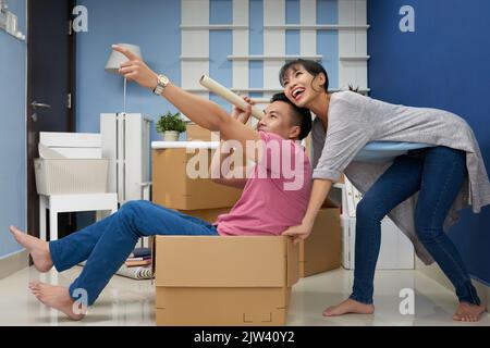 Junges glückliches asiatisches Paar, das sich in einer neuen Wohnung unterhielt, Mann, der in einem Karton sitzt und in einem Stretchfilmteleskop schaut Stockfoto