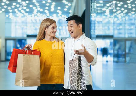 Junge glückliche verheiratete Paar Käufer Mann und Frau im Supermarkt sind glücklich und lachen miteinander, reden mit Einkaufstaschen und Geschenken. Stockfoto