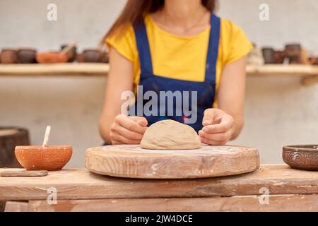 Frau in blauer Schürze und gelbem T-Shirt arbeitet auf mechanischem Töpferrad vor dem Hintergrund von Regalen mit Tonprodukten Stockfoto