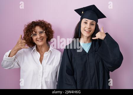 Die hispanische Mutter und Tochter tragen eine Abschlusskappe und ein Zeremoniengewand und lächeln beim Telefonieren mit Händen und Fingern, als ob sie am Telefon sprechen würden. Stockfoto