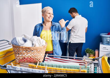 Hispanische Mutter und Sohn hängen Kleider an der Wäscheleine und zeigen den Daumen nach oben zur Seite, lächelnd mit offenem Mund Stockfoto