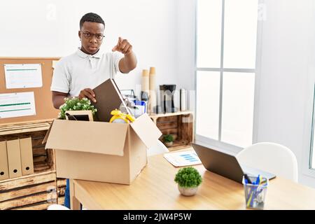 Junger afrikanischer Mann, der Büroobjekte in einen Karton legt und mit dem Finger auf die Kamera und auf Sie zeigt, selbstbewusste Geste, die ernst aussieht Stockfoto