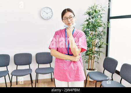 Junge asiatische Krankenschwester Frau im medizinischen Wartezimmer schaut zuversichtlich auf die Kamera lächelnd mit gekreuzten Armen und Hand auf dem Kinn angehoben. Denken positiv. Stockfoto