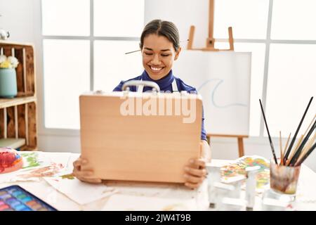 Junge latinerin lächelt selbstbewusst und eröffnet eine Aktentasche im Kunststudio Stockfoto