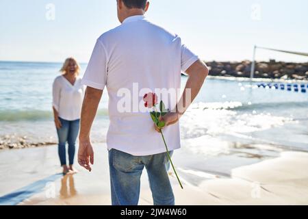 Mann und Frau im mittleren Alter überraschen am Meer mit Blumen auf dem Rücken Stockfoto