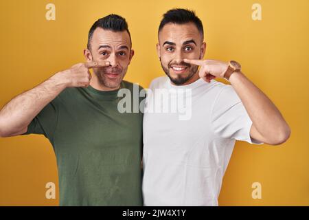 Homosexuelles Paar, das auf gelbem Hintergrund steht und mit dem Finger auf Gesicht und Nase zeigt, lächelt fröhlich. Schönheitskonzept Stockfoto