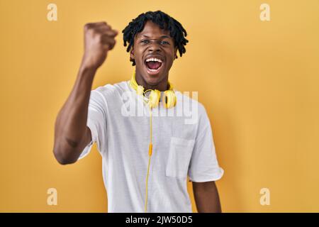 Junger afrikanischer Mann mit Dreadlocks, die auf gelbem Hintergrund stehen, wütend und wütend, die Faust zu erheben, frustriert und wütend, während er vor Wut schreit Stockfoto