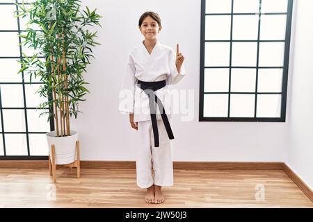 Junge hispanische Mädchen trägt Karate Kimono und schwarzen Gürtel zeigt und zeigt mit Finger Nummer eins, während lächelnd zuversichtlich und glücklich. Stockfoto