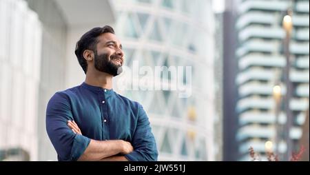 Glücklicher reicher indischer Geschäftsmann, der in der Großstadt steht und an Zukunftsvisionen denkt. Stockfoto