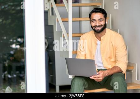 Lächelnder indischer Mann, der mit einem Laptop auf der Treppe sitzt. Hochformat Stockfoto