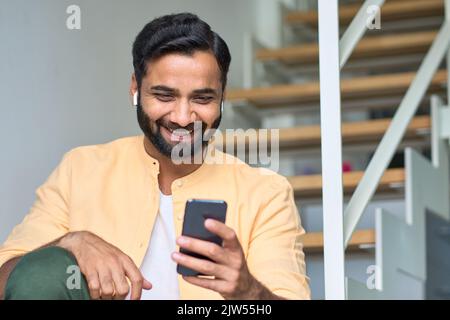 Indischer Mann, der zu Hause mit Ohrstöpseln sitzt und Videos mit dem Mobiltelefon ansieht. Stockfoto