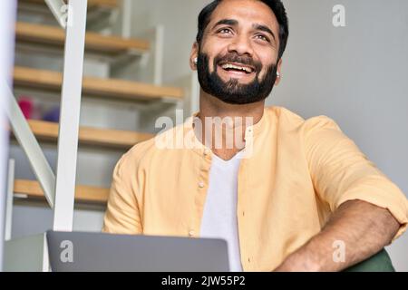 Glücklich lachende ethnische indische Geschäftsmann trägt Ohrstöpsel mit Laptop. Stockfoto