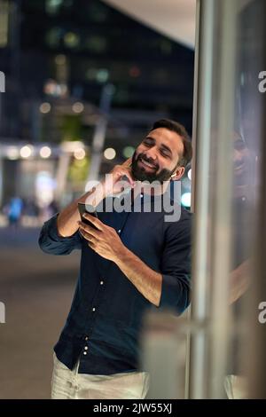 Ein indischer Mann mit Ohrstöpseln hält das Telefon und hört nachts mobile Musik. Stockfoto