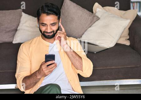 Indischer Mann sitzt zu Hause auf dem Sofa mit dem Handy und schaut sich Videos an. Stockfoto