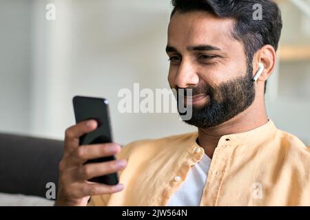 Lächelnder indischer Mann, der zu Hause mit Kopfhörern per Smartphone sitzt. Stockfoto