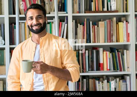 Glücklicher indischer Mann, der in der Nähe des Bücherregals mit Ohrstöpseln steht und Hörbuch hört. Stockfoto