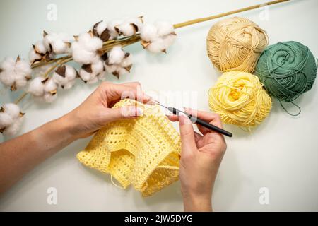 Die Hände der Frau stricken Babykleidung aus gelbem Garn, auf einem weißen Tisch, Draufsicht. Stockfoto