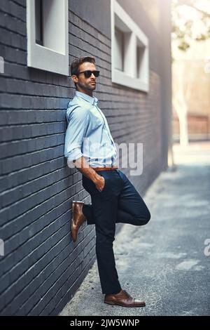 Er macht Geschäfte mit Stil. Ein hübscher junger Geschäftsmann, der gegen eine graue Backsteinmauer steht. Stockfoto