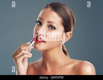 Nichts sagt kühn ganz wie rote Lippen. Studioportrait einer schönen jungen Frau, die auf einem grauen Hintergrund einen roten Lippenstift aufsetzt. Stockfoto