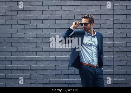 Erfolg geht nie aus dem Stil. Ein hübscher junger Geschäftsmann steht gegen eine graue Backsteinmauer. Stockfoto