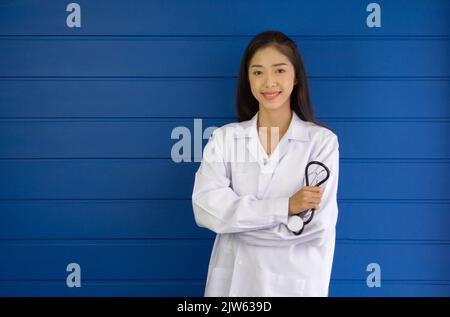 Junger asiatischer Arzt in weißem Kleid und Stethoskop mit Armkreuz vor blauer Wand. Stockfoto