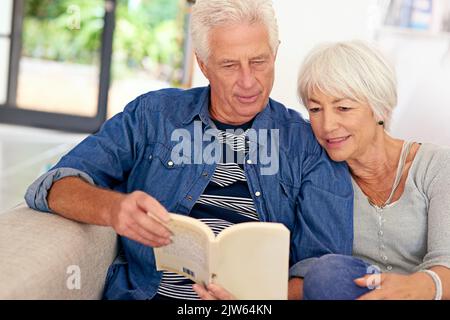 Ein entspanntes Seniorenpaar, das zu Hause auf dem Sofa ein Buch liest, lebt ihre ganz eigene Liebesgeschichte. Stockfoto