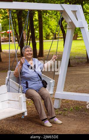 Verspielte und glückliche Frau genießen bei Schaukel im Freien in Park oder Garten entspannt lachen, alte Menschen freudig aktiven Ruhestand und Entspannungskonzept. Stockfoto