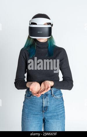 Ein Mädchen im Teenageralter mit blauen Haaren, das eine Virtual-Reality-Brille trägt, Filme gucken oder Videospiele spielt, hält ihre leeren Handflächen zusammen, als ob sie nach ihr fragen würde Stockfoto