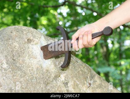 Hand des Ritters, der versucht, das Excalibur-Schwert zu extrahieren, das in den Felsen inmitten des verzauberten Waldes eingebettet ist Stockfoto