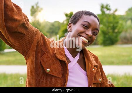 Porträt einer selbstbewussten, fröhlichen afroamerikanischen jungen Frau mit kurzen Haaren, unbeschwert im Freien in der Parklandschaft, mit offenen Armen, lächelnd und schauend auf Th Stockfoto