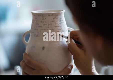 Weibliche Kunsthandwerkerin malt in der Werkstatt Text auf weiße Keramikvase mit Marker auf verschwommenem Hintergrund Stockfoto